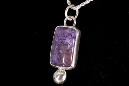 Purple Charoite pendant on a silver chain against a black background right side view